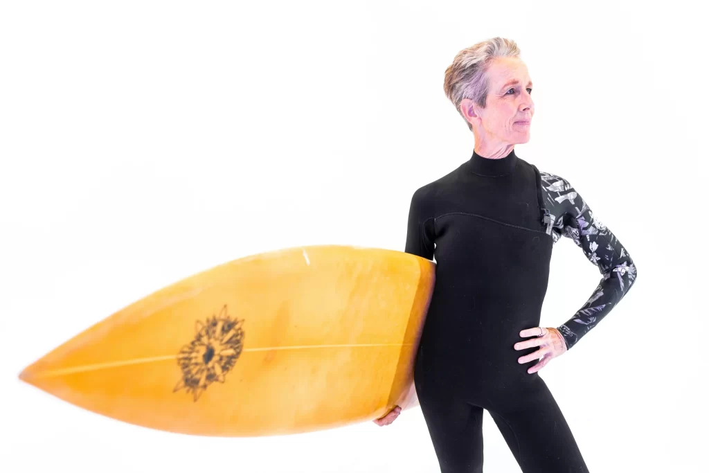 An elderly lady with grey and pink hair in a wetsuit holding a surfboard.